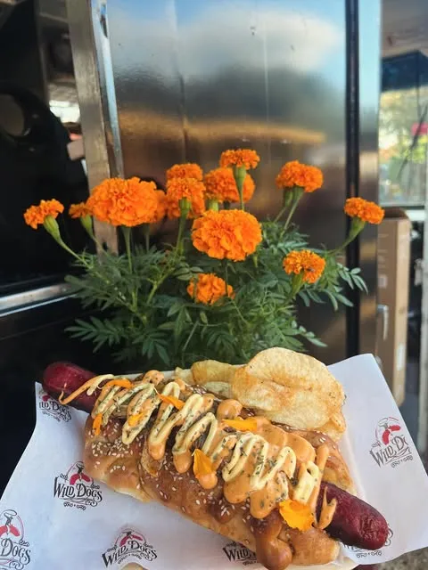 Hot dog con cebolla asada, papitas y aderezos, junto a flores de cempasúchil